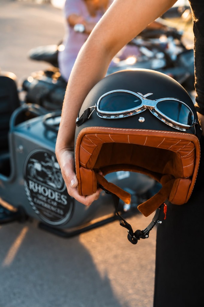 A guest holding a vintage leather helmet with goggles beside a Rhodes sidecar